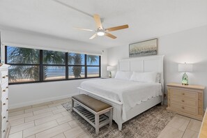A bedroom with a bed, a bench, a nightstand, a lamp, a ceiling fan, and a window with a view of palm trees.