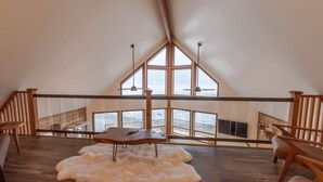 Une pièce mansardée spacieuse avec un plafond en pente, un plancher en bois, un tapis blanc et moelleux, une table en bois et une vue sur un balcon doté d’une balustrade.