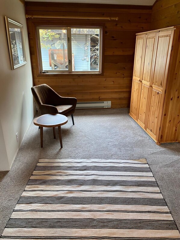 A room with a wooden floor, a striped rug, a round table, a brown chair, and a wooden wardrobe.