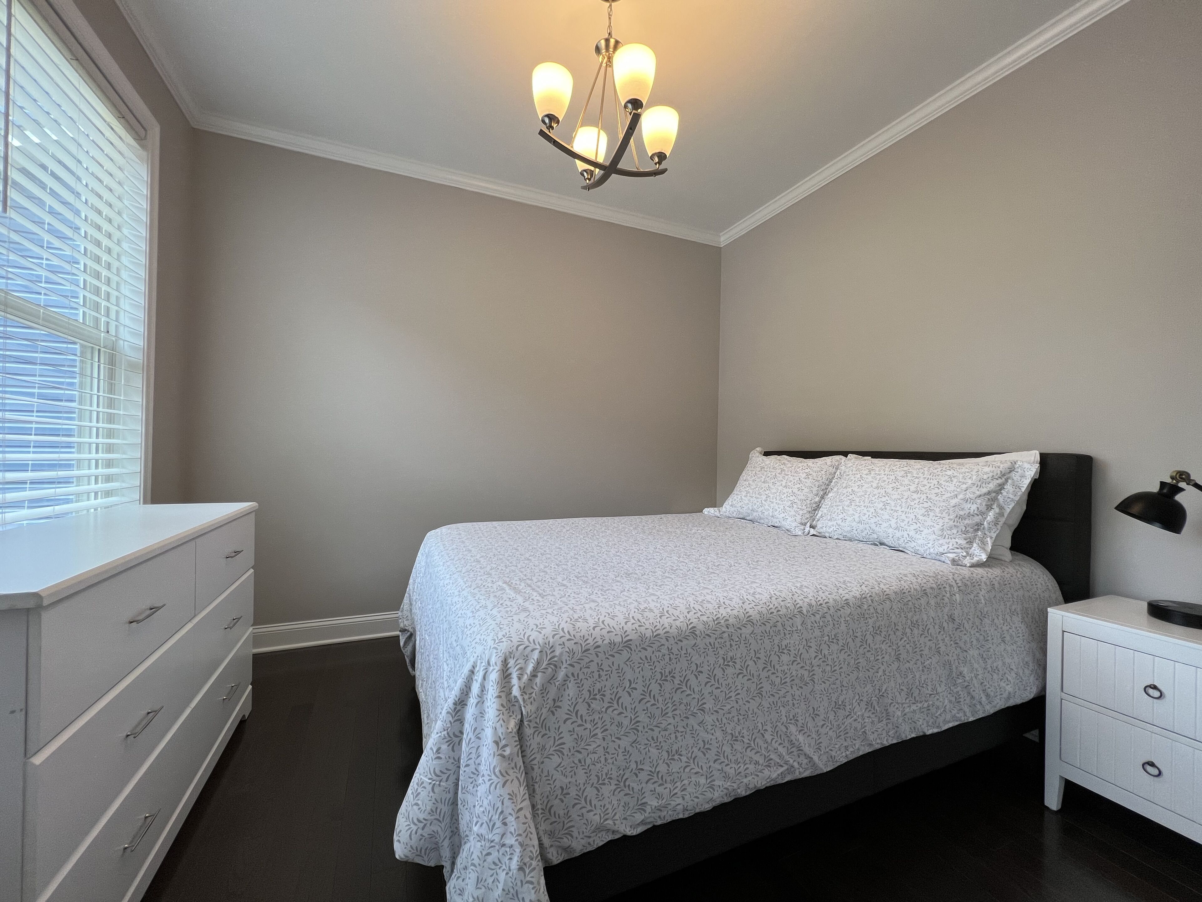 A bedroom with a bed, a nightstand, a dresser, a window with blinds, and a chandelier.