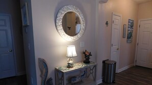 Un couloir avec un miroir rond, une table avec une lampe et une poubelle.