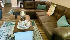 Un salon avec un canapé en cuir marron, une table basse agrémentée d’objets décoratifs, un tapis à motifs et, au fond, une cuisine.