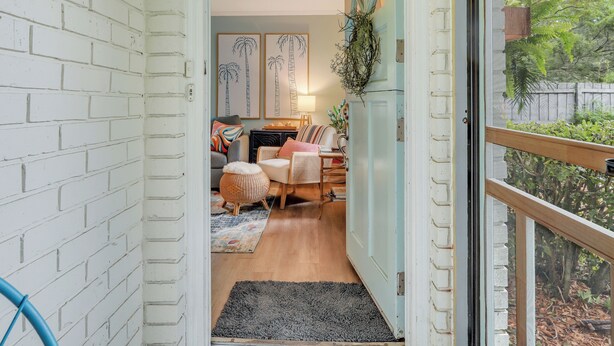 A cozy room with a white brick wall, a wooden floor, a black rug, a wicker chair, a sofa, and a small table with a lamp.