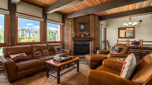 A cozy living room with leather sofas, a fireplace, and large windows offering a view of a building and trees. - Downhill Den! Mountain Views, Private Patios, Outdoor Pool, 4 Hot Tubs, Pickleball Courts! (Steamboat Springs)