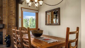 A wooden dining table with chairs, a woven basket, and a placemat. A fireplace is visible to the left, and a window with a view of trees is on the right. - Downhill Den! Mountain Views, Private Patios, Outdoor Pool, 4 Hot Tubs, Pickleball Courts! (Steamboat Springs)