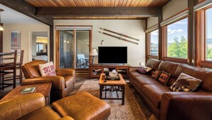 A cozy living room with leather furniture, a television, and large windows offering a view of the outdoors. - Downhill Den! Mountain Views, Private Patios, Outdoor Pool, 4 Hot Tubs, Pickleball Courts! (Steamboat Springs)