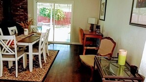 A dining area with a wooden table and chairs, a small side table with a lamp, and a view of the outdoors through a glass door.