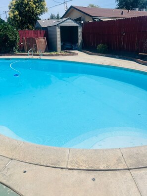 Une piscine avec une plage en béton, entourée d’une clôture en bois et d’un abri de jardin.