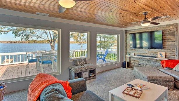 A spacious living area with large windows overlooking a lake, a ceiling fan, and a flat-screen TV mounted on the wall. - Dock & Fire Pit: Lakefront Cedar Creek Home! (Mabank)