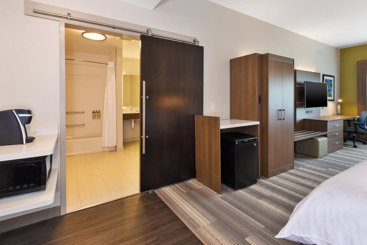 Ein Hotelzimmer mit einer Schiebetür zum Badezimmer, einem Schreibtisch mit Stuhl, einem Bett und einem an der Wand befestigten Fernseher.
