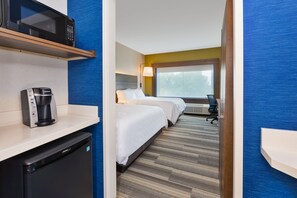 Ett hotellrum med mikrovågsugn, kaffebryggare och diskmaskin på bänken, två sängar med vita sängkläder, ett skrivbord med en lampa och ett stort fönster med utsikt över träd.