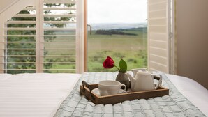 Ein Schlafzimmer mit einem Bett, einem Tablett mit Teekanne, Tasse und einer Rose, sowie Blick auf eine grüne Landschaft durch geöffnete Fensterläden.