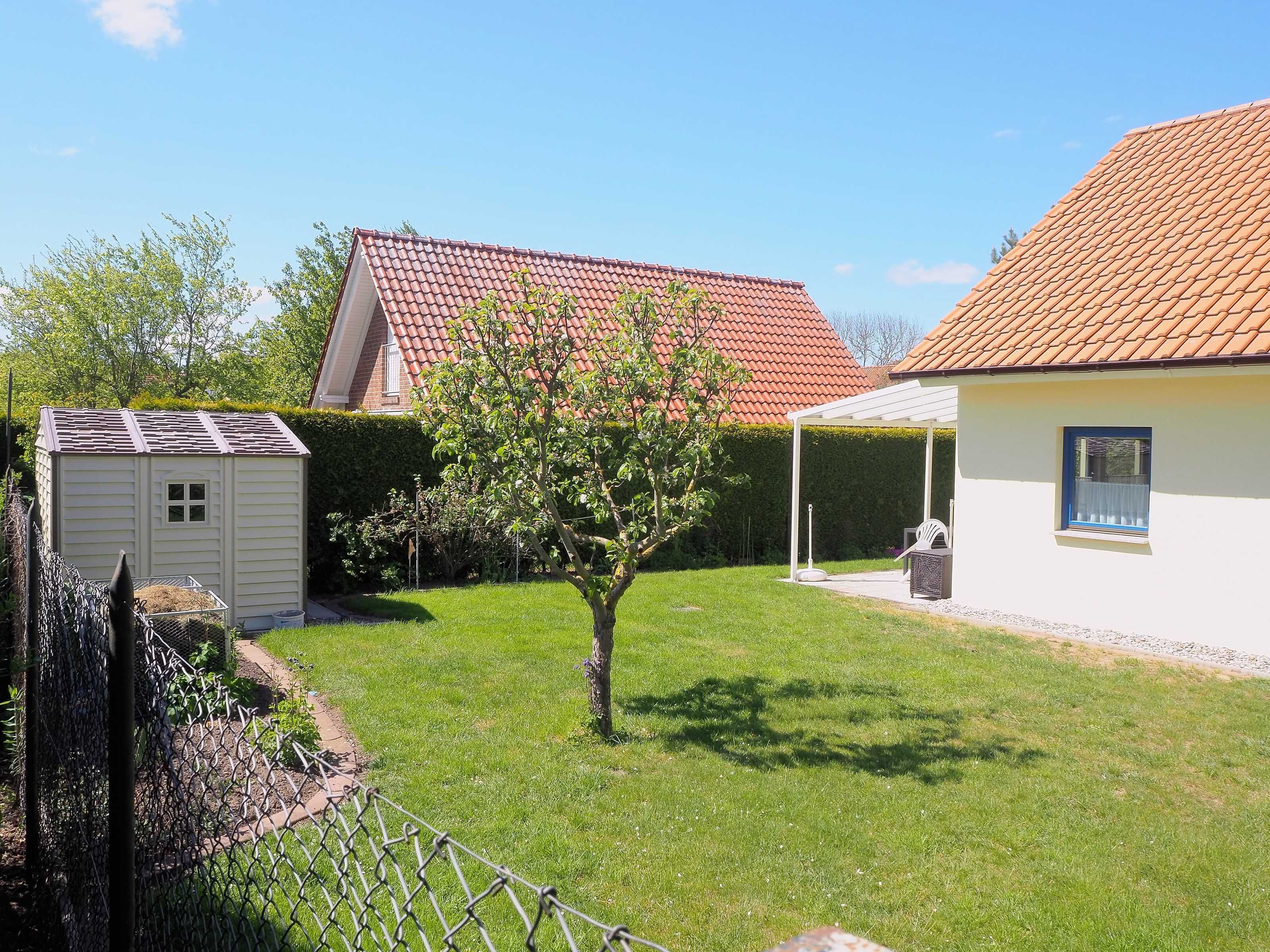 Ein Wohnhaus mit eingezäuntem Garten, einer kleinen Gartenhütte, einem Baum und einem Dach mit roten Ziegeln.