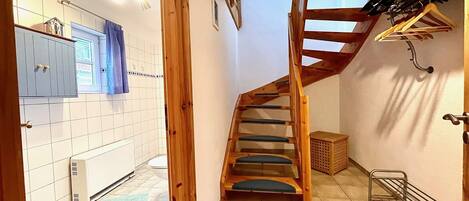 Un escalier menant à une chambre à coucher située en mezzanine, avec un plafond en bois, une cuisine dotée de meubles blancs et un porte-chaussures suspendu.