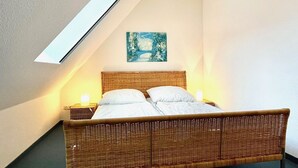 Una habitación con techo inclinado, una cama con sábanas blancas, una mesita de noche con lámpara y un cuadro en la pared.