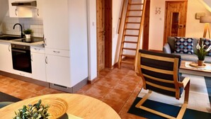 Cocina compacta con armarios blancos, horno integrado y un lavamanos. Una escalera de madera conduce a una zona de dormir en un altillo con una cama y un sofá.