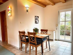 A dining area with a wooden table and chairs, a potted plant, a wall-mounted light fixture, and a window with a view of greenery.