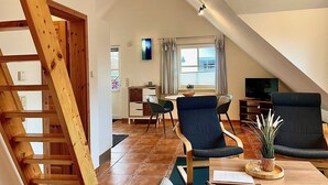 A cozy loft-style living room with a wooden staircase, a dining area, and a TV. - Ferienwohnung Haus 3 Wohnung 2 - Ferienhäuser Liethmann (Insel Poel)