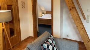 A cozy room with a wooden ceiling, a sofa with a patterned cushion, and a bed in the background. - Ferienwohnung Haus 3 Wohnung 2 - Ferienhäuser Liethmann (Insel Poel)