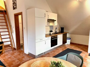Une cuisine compacte avec des meubles blancs, une cuisinière et un lave-vaisselle. Contre le mur, on aperçoit une échelle en bois et, près de là, une petite table sur laquelle est posée une plante.