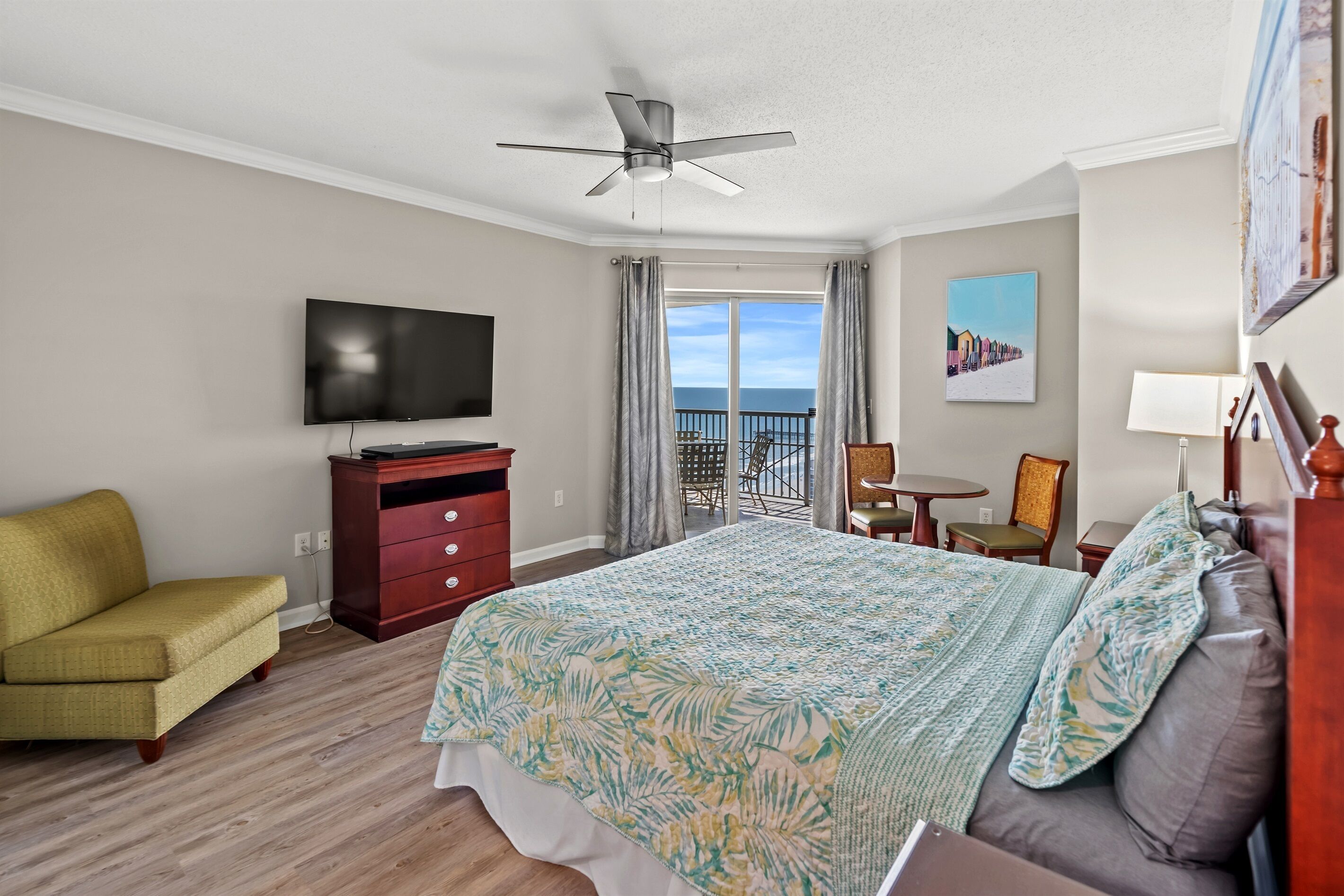 A hotel room with a bed, a television, a desk, a chair, a sofa, and a balcony with a view of the ocean.