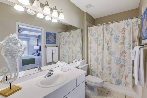 Un baño con un lavabo blanco, un inodoro, ducha con cortinas florales, un espejo grande y una lámpara en el techo.