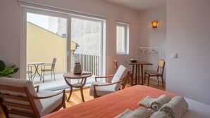 Ett hotellrum med en säng, två stolar, ett litet bord, en lampa och en balkong med ett bord och stolar.