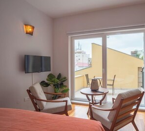 A hotel room with a large sliding glass door to the balcony, a flat-screen TV mounted on the wall, and a dining area with a table and chairs.