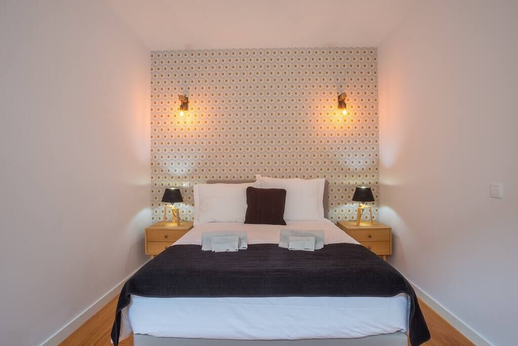 A neatly made bed with a dark blanket, two pillows, and two towels on the side. There are two bedside tables with lamps and a patterned wallpaper on the wall behind the bed.
