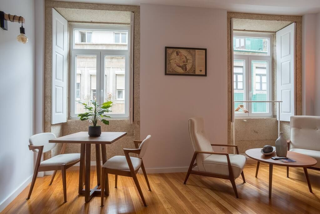 Quarto com piso de madeira, uma mesa de jantar com cadeiras, um vaso de planta e duas janelas com vista para prédios.