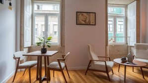 Una habitación con piso de madera, una mesa de comedor con sillas, una planta en maceta y dos ventanas con vistas a edificios.