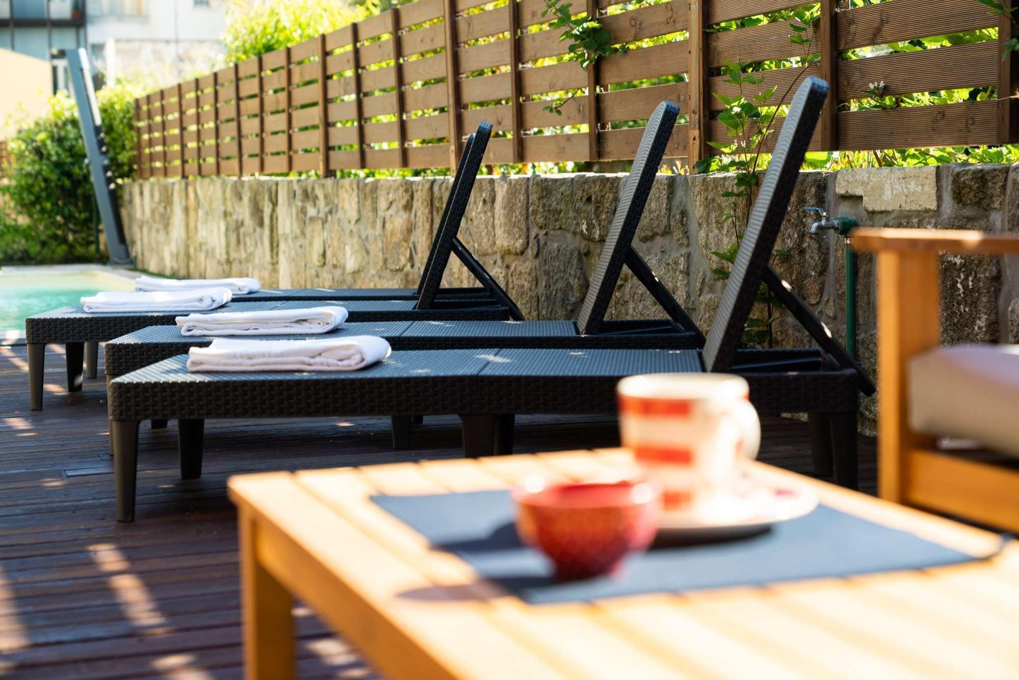 Un espace extérieur ensoleillé, aménagé avec des chaises longues et une table en bois, des serviettes soigneusement pliées et une tasse posée sur la table.