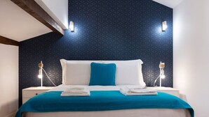 Una cama bien tendida con una colcha color turquesa y almohadas blancas, dos lámparas de noche y una pared oscura con estampado.