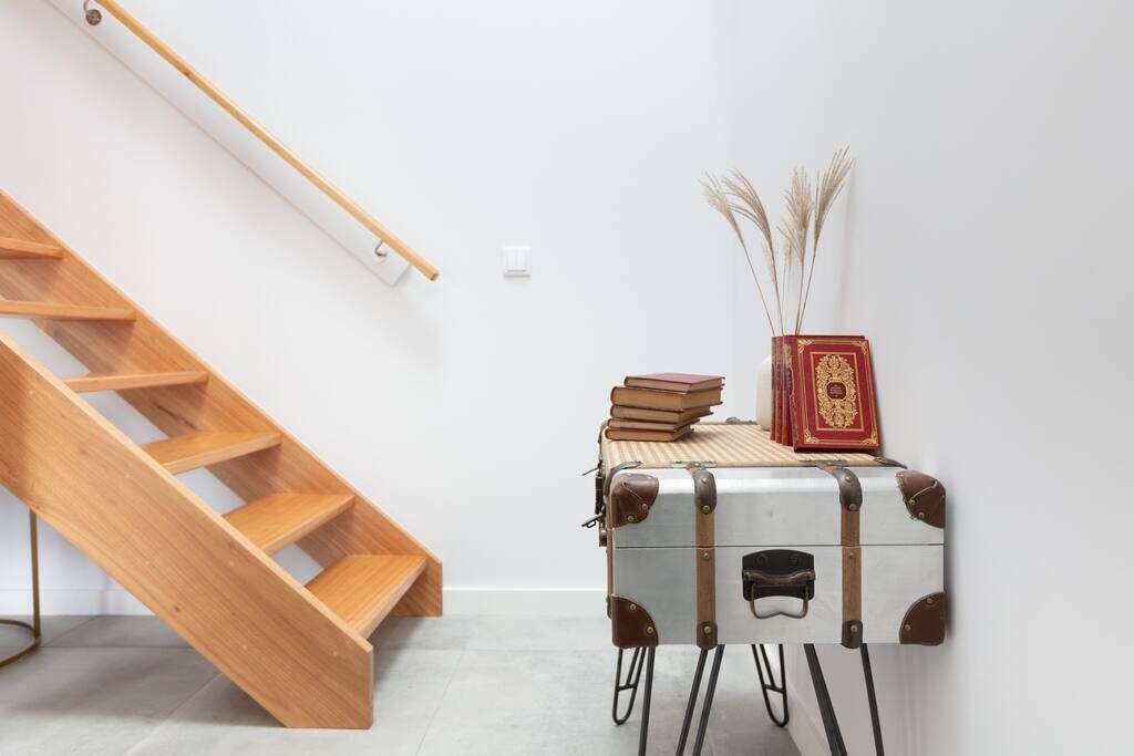 Una scala in legno addossata a un muro bianco, una valigia vintage con libri e oggetti decorativi sopra, e un tavolino con un vaso di piante essiccate.