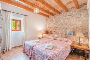 A bedroom with two beds, a stone wall, a wooden ceiling, and a window with curtains. - Beautiful Vacation Home "Can Faveta" with Mountain View, Pool, Wi-Fi (Alcudia)