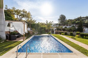Una piscina rectangular con superficie de agua azul cristalina, rodeada por una terraza pavimentada, una zona de estar cubierta con cojines y un jardín bien cuidado con diversas plantas y césped.
