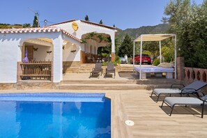 Un área junto a la piscina con sillones reclinables, una zona de estar techada y un edificio con antena parabólica.