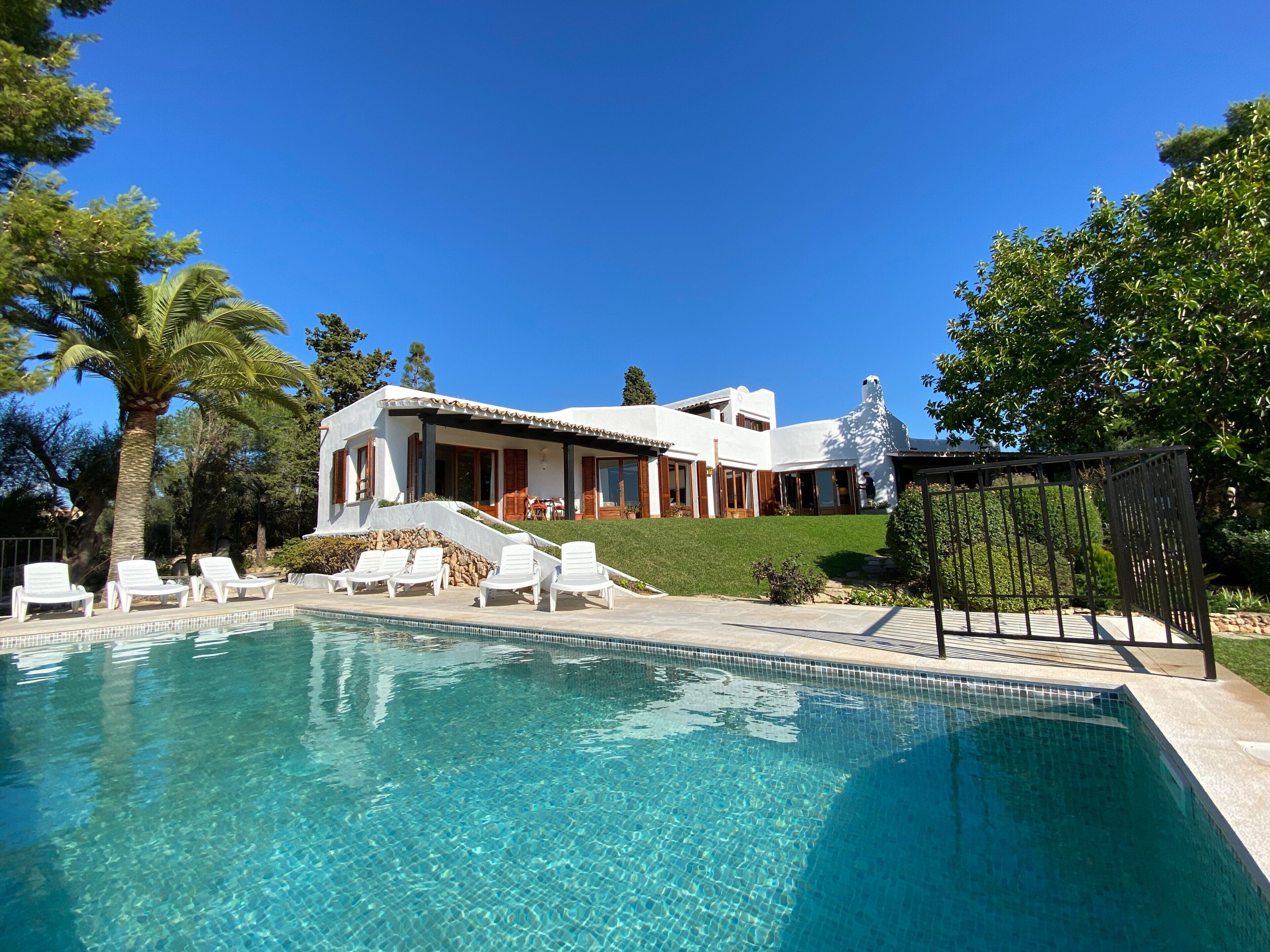 Piscina com espreguiçadeiras, um edifício branco com telhado inclinado e um céu azul límpido.