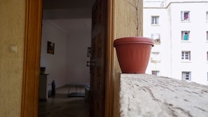 Een binnengang met een houten deur die naar een kamer leidt, een bloempot op een balkon en een gebouw met balkons op de achtergrond.