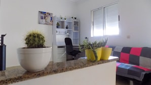 Een woonkamer met een bank, een bijzettafeltje en een cactus in een pot. Er is een raam met zonwering en een schilderij aan de muur.