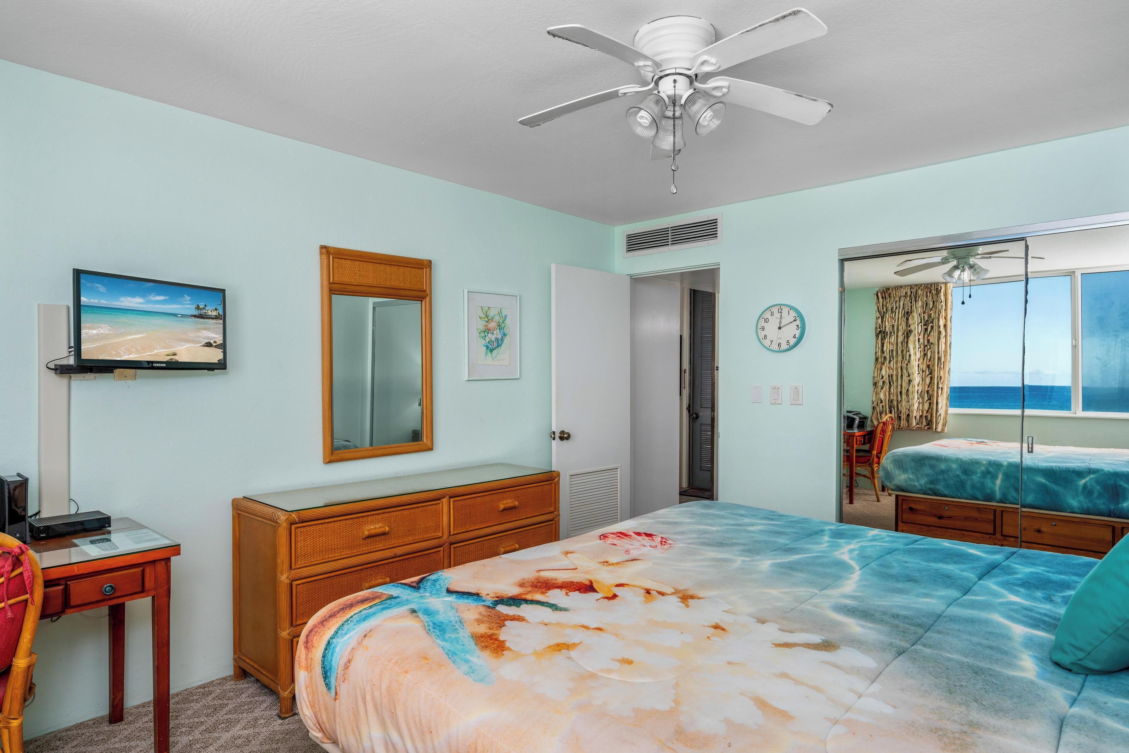 Un dormitorio con cama, escritorio, cómoda, ventilador de techo, ventana con cortinas y vista al océano.