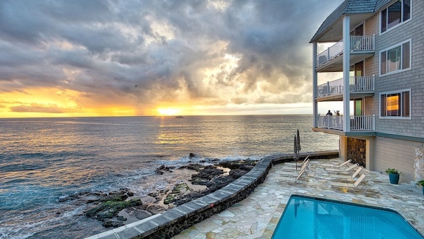 A pool area with a view of the ocean at sunset, a stone deck, and a building with balconies.