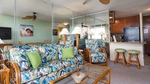 A living room with a floral sofa, wooden chairs, a glass coffee table, and a kitchen area with wooden cabinets and a refrigerator.