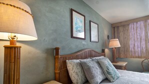 A bedroom with a wooden headboard, a bed with patterned bedding, two framed pictures on the wall, and a window with vertical blinds.