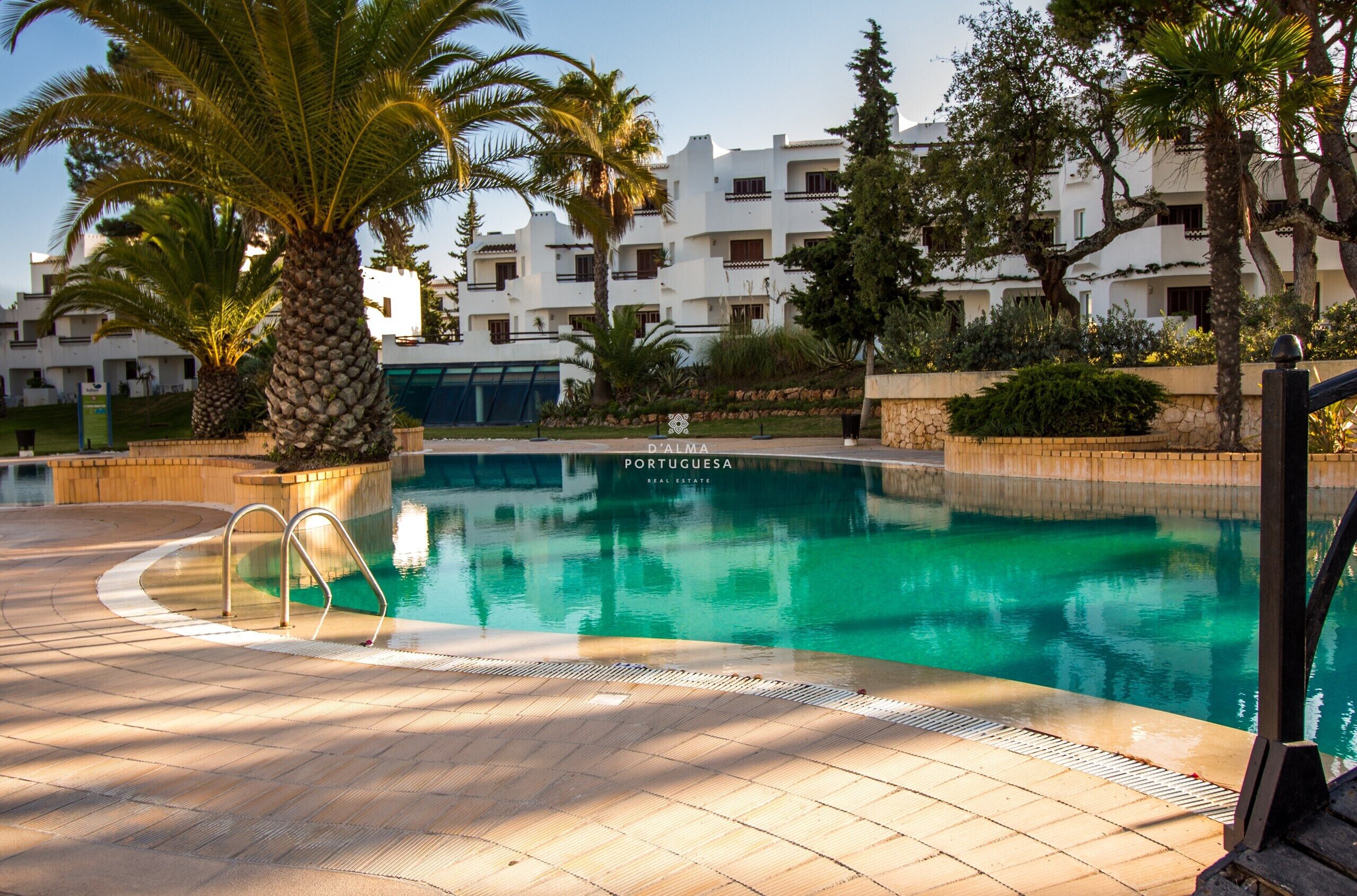 A swimming pool with a tiled deck, palm trees, and a building with balconies in the background.