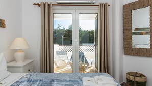 Una habitación de hotel con cama, mesita de noche, lámpara, ventana con cortinas, balcón con sillas y espejo.
