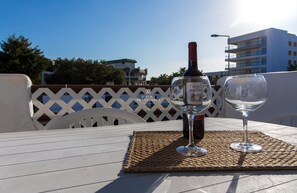 Une bouteille de vin et un verre vide sont posés sur une table recouverte d’un set de table en fibres tressées, le tout se détachant sur un fond constitué d’une clôture en treillis blanc et de bâtiments en arrière-plan.