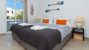 Un dormitorio con cama, dos tablas de remo fijadas a la pared, un cojín con motivos playeros, una lámpara y vista a un edificio a través de un ventanal.