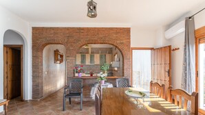A dining area with a brick archway, wooden chairs, a wooden table, and a kitchen in the background. - Holiday Home "Casa Rural Villena" with Sea View, Wi-Fi, Pool & Terrace (Frigiliana)