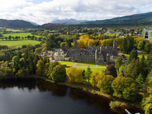 A large, historic building with multiple towers and a well-maintained lawn, surrounded by lush greenery and a river.